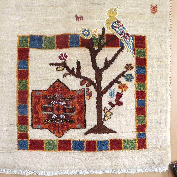 色鮮やかな鳥と木のモチーフが描かれた手織りのウール製ギャッベラグが、ペルシャ絨毯専門店の通販商品として床に広がっている。