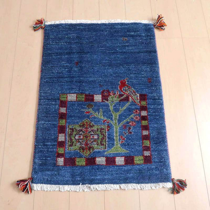 ギャッベ／ルリバフ 84×61cm（HSS-71836）は、鮮やかな青色のウール製ギャッベラグが木製の床に敷かれ、ペルシャ絨毯のような手織りの模様と鳥のデザインが際立っている。