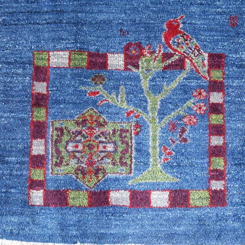 青い背景に赤い鳥と木のデザインが丁寧に手織りされたギャッベのラグが見える。