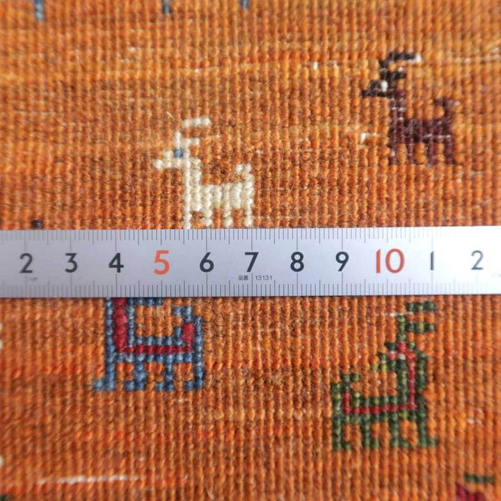 オレンジ色の手織りギャッベラグに、さまざまな色の動物模様が施され、ウールの温かみが定規越しに感じられる。