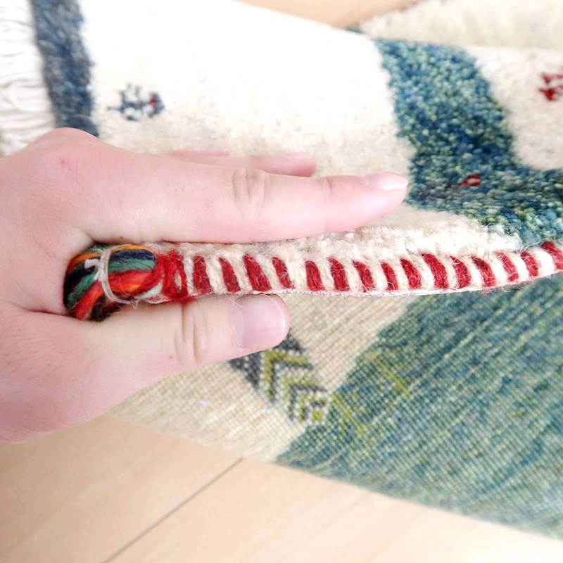 手織りのギャッベラグが指で持ち上げられ、ウールの厚みと柔らかい質感が際立っています。