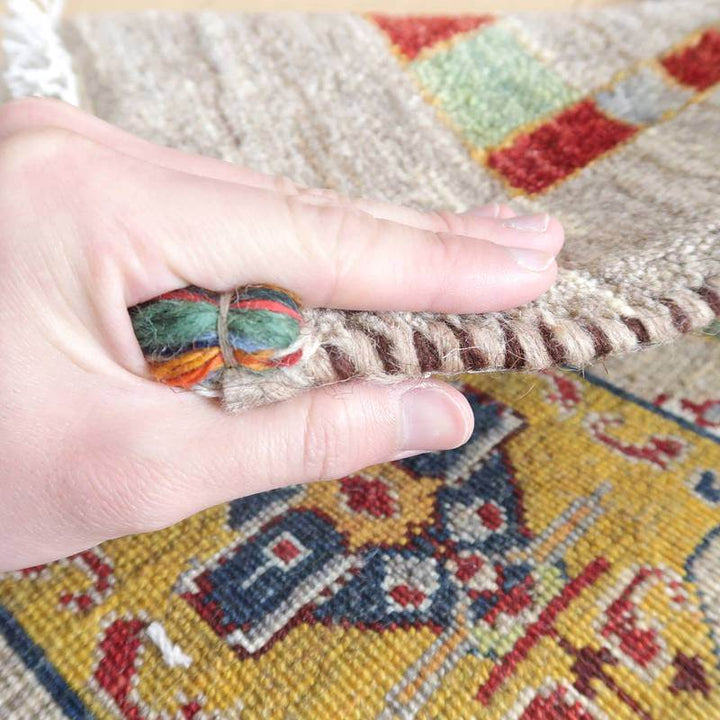 手織りのギャッベラグを指でつまんで厚みを確認している様子で、ウール素材の柔らかさとペルシャ絨毯のような豊かな色彩が際立っている。