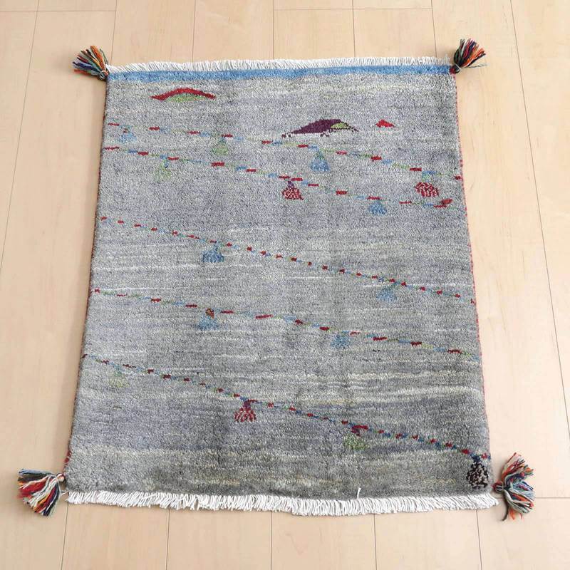 ギャッベ／ルリバフ 75×64cm（HSS-72423）は、グレーの手織りギャッベラグが木製フローリングの上に広がり、色とりどりのタッセルがアクセントとなっている。
