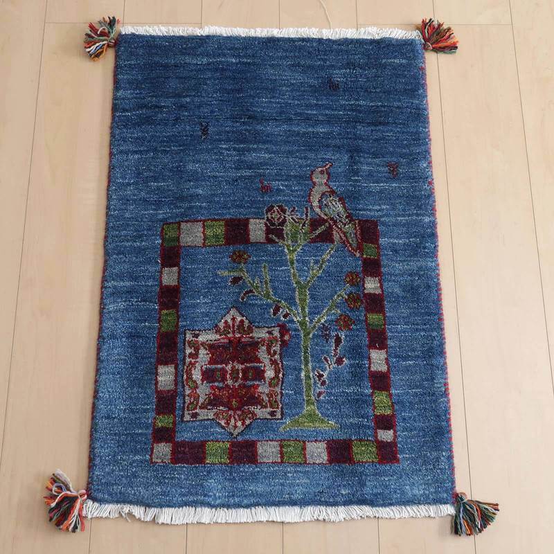 ギャッベ／ルリバフ 86×59cm（HSS-72487）は、青を基調に鳥と木のデザインが施された手織りウールのギャッベラグが、木目の床に美しく敷かれている。