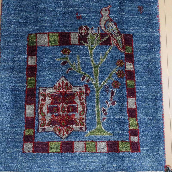 青い背景に鳥と木のデザインが描かれた手織りのギャッベラグが床に敷かれている。