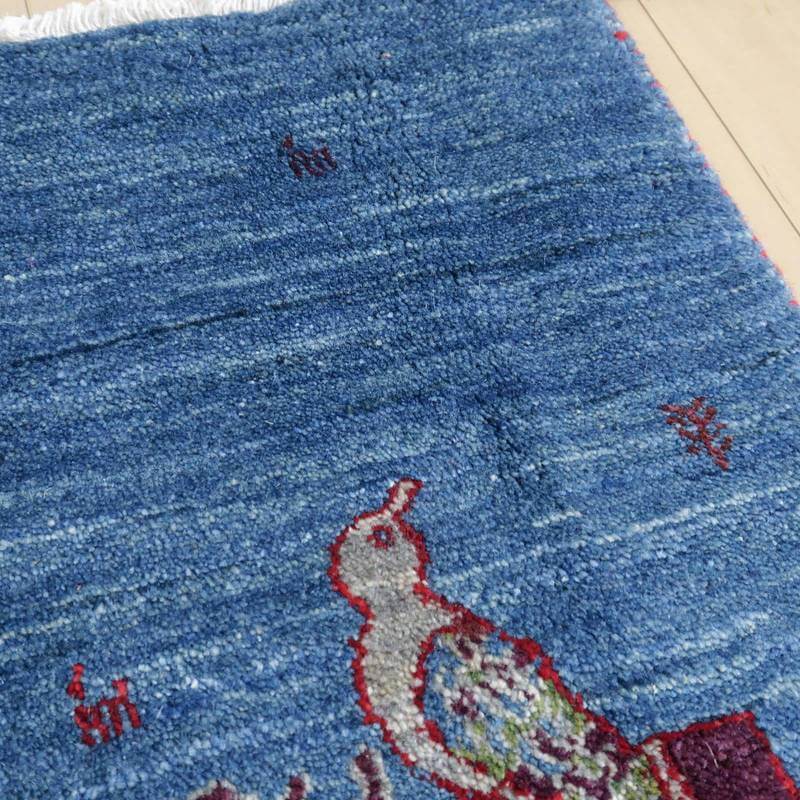 青いウールの手織りギャッベラグに描かれた鳥の模様が、ペルシャ絨毯専門店の通販商品として床に映えている。