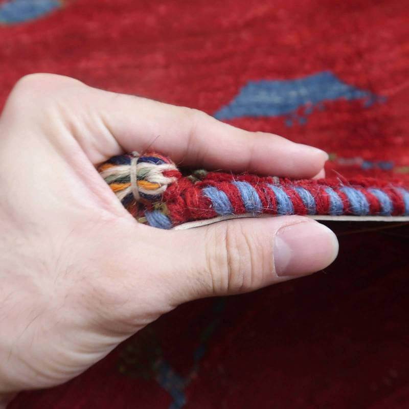 手で持たれたギャッベの厚みが見える接写画像で、手織りウールの色鮮やかな模様が際立ち、通販専門店で購入できるペルシャ絨毯の質感が伝わる。