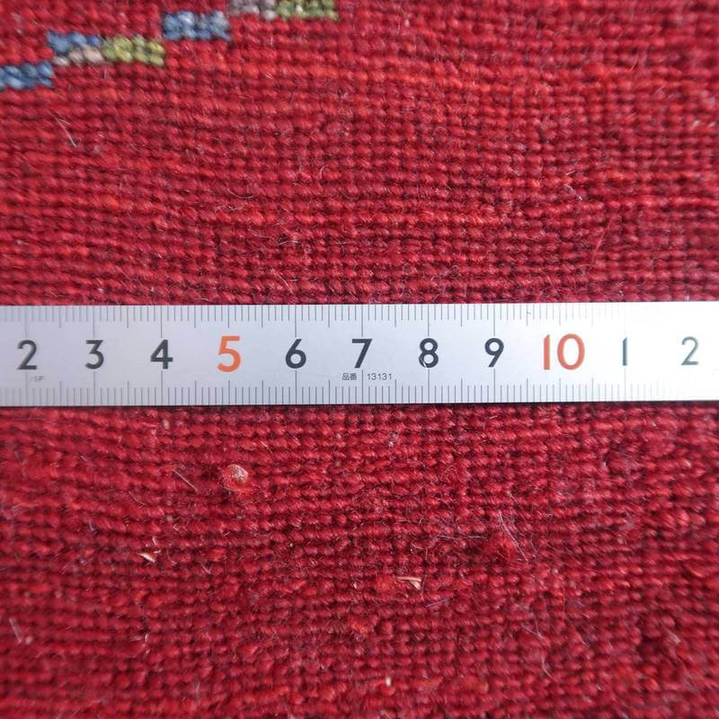 赤い手織りのウールギャッベラグの細やかな織り目と定規が並び、ペルシャ絨毯専門店の通販で購入可能な質感が際立っている。