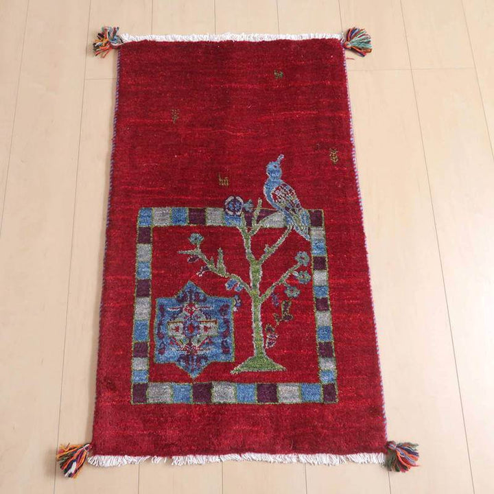 ギャッベ／ルリバフ 90×55cm（HSS-72565）は、赤いウールのギャッベラグが、木目の床に鮮やかに敷かれ、ペルシャ絨毯専門店の通販で手織りの温もりが際立っている。