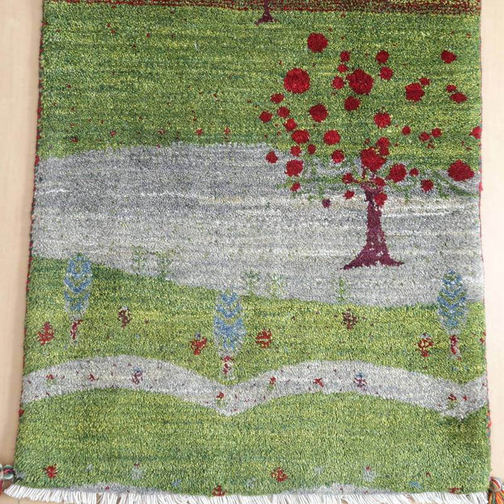 緑の草原に赤い木々が描かれた手織りのギャッベラグは、ウール素材特有の温かみがあり、ペルシャ絨毯専門店の通販で購入できる逸品が際立っている。