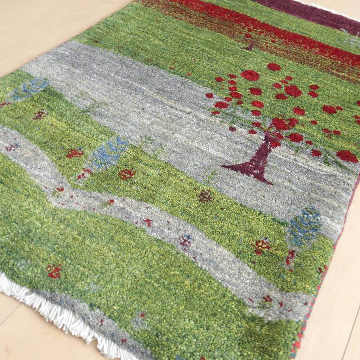 緑豊かな風景と赤い木が描かれた手織りのギャッベラグが床に敷かれ、ウールの温もりが伝わるペルシャ絨毯の専門店通販品が見える。