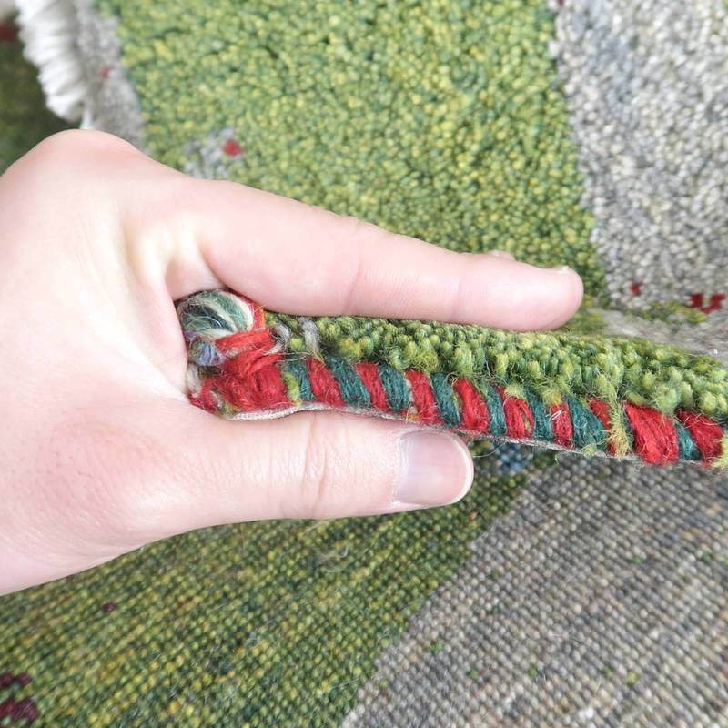 手で持たれたギャッベのラグが、手織りウールの厚みと色鮮やかなペルシャ絨毯の縁取りを際立たせている。