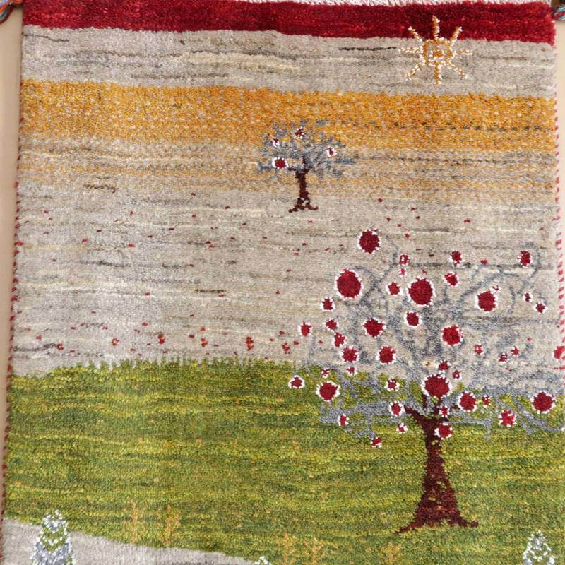 緑の草原に赤い花が咲く木が描かれた手織りのギャッベラグが、暖かいウールの質感とともにペルシャ絨毯専門店で通販されています。