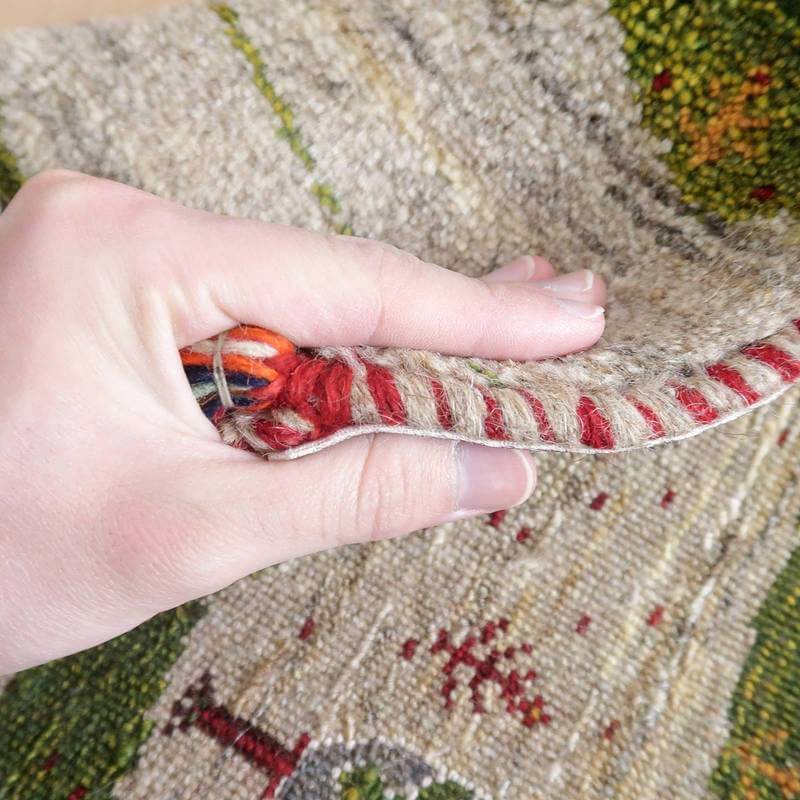 手に取られたギャッベのラグが、丁寧に手織りされたウールの温かみを伝えており、赤と緑の模様が織り目の細かさを強調している。