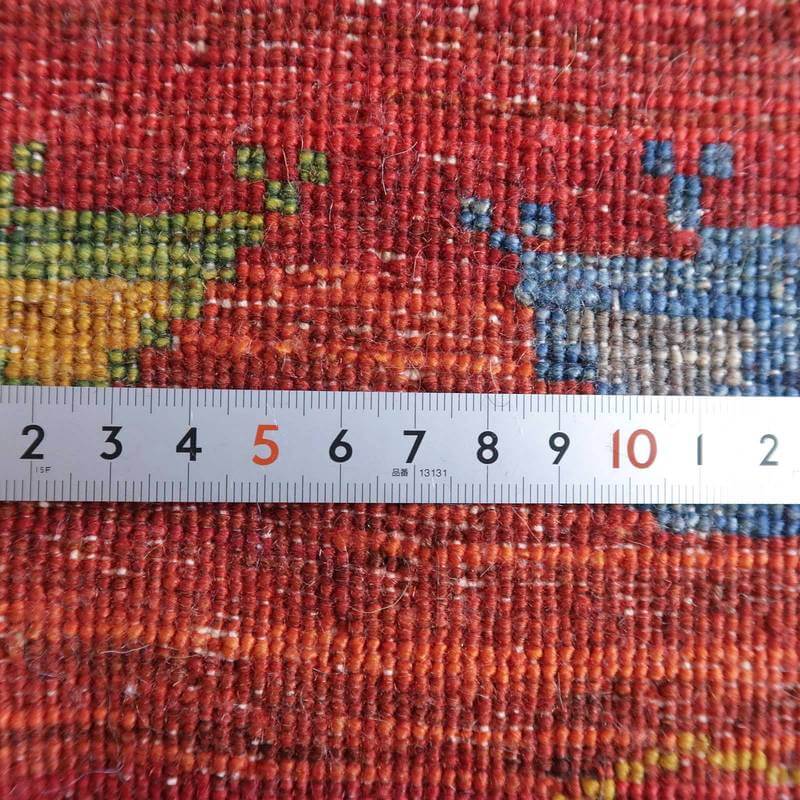 赤と青の模様が鮮やかに映える手織りのギャッベラグに定規が置かれ、ウールの織り目の密度が際立っている。