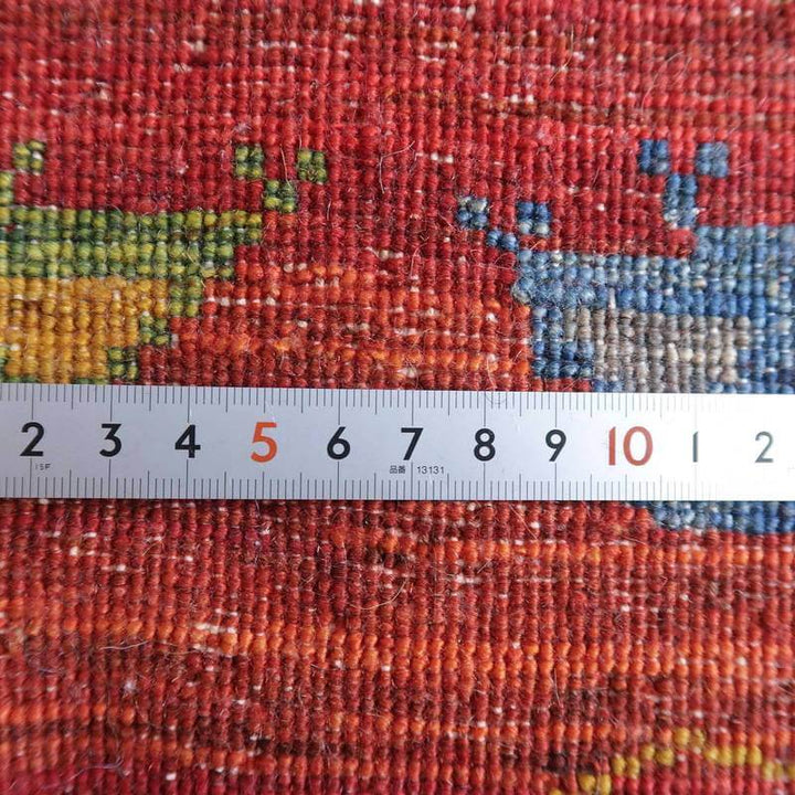 赤と青の模様が鮮やかに映える手織りのギャッベラグに定規が置かれ、ウールの織り目の密度が際立っている。