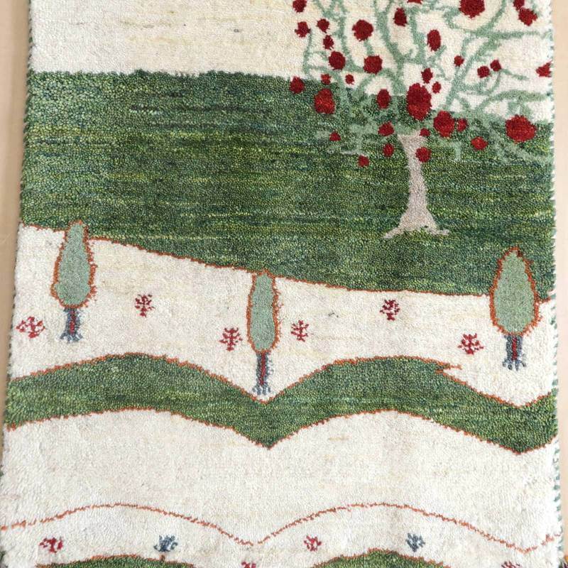 手織りの温もりが伝わるウール素材のギャッベラグには、緑豊かな風景と赤い花が描かれており、専門店の通販で個性的なペルシャ絨毯が見える。
