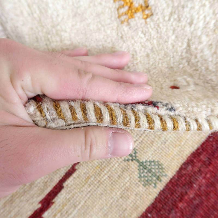 手織りのギャッベラグが指で持ち上げられ、ウールのしっかりした厚みと温かみが感じられる質感が際立っています。