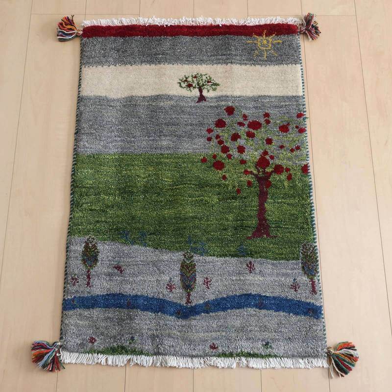 緑と赤の木々が描かれた手織りのウール製ギャッベラグが、ペルシャ絨毯専門店の通販で床に広げられている。