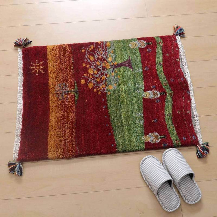 赤と緑の色彩が鮮やかな手織りのギャッベラグが、木の模様と共にぬくもりを伝え、ペルシャ絨毯専門店の通販で見つけた一品が床に敷かれている。