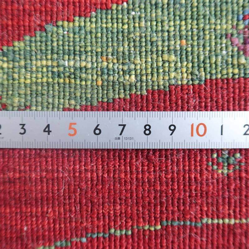 赤と緑の色合いが美しい手織りウールのギャッベラグは、ペルシャ絨毯専門店の通販で購入可能で、織りの密度が定規で詳細に測定されている。