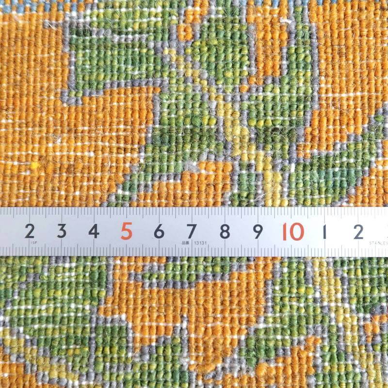 オレンジとグリーンの模様が鮮やかに織り込まれた手織りウールのギャッベラグが、定規との比較で細やかな織り目が際立っている。