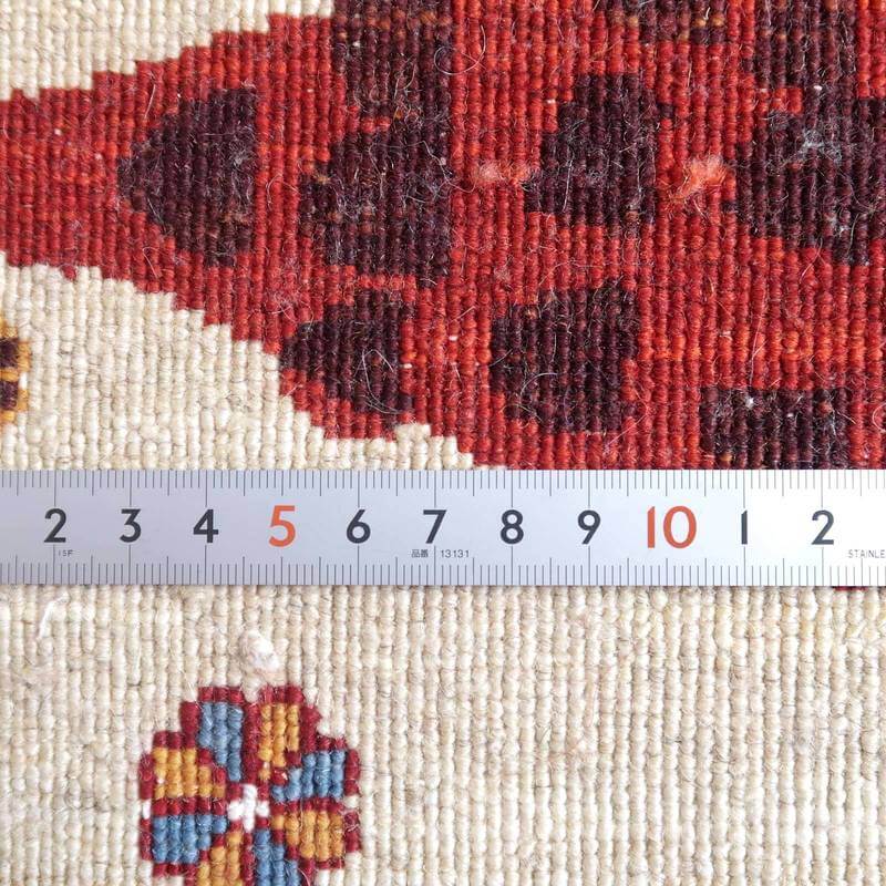 赤とクリーム色の模様が美しい手織りのギャッベラグが、定規とともに映し出され、繊細なウールの織り目が際立っている。