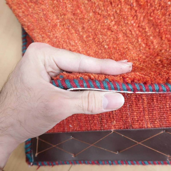 オレンジとブルーの縁取りが美しい手織りウールのギャッベラグの厚みが指に挟まれ、ペルシャ絨毯専門店の通販で見られる質感が感じられる。