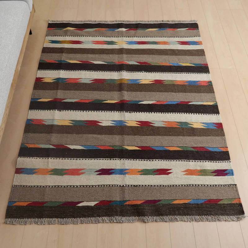 キリム・シラーズ／カシュガイ 203×154cm（KL-796）は、色とりどりのストライプ模様が特徴の手織りウール製ギャッベラグが、木目の床にぴったりと敷かれている。
