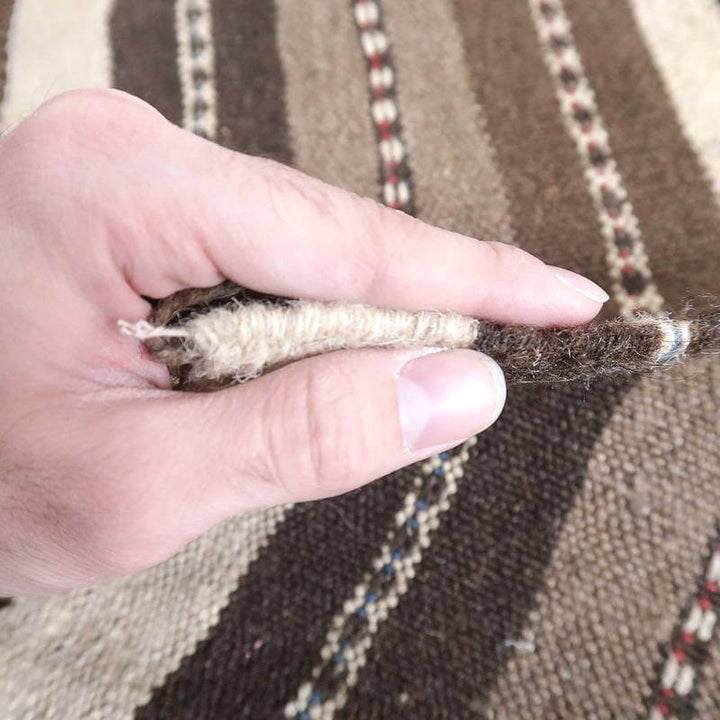手織りのギャッベラグが指で挟まれ、ウールの厚みと織りの密度が際立ち、ペルシャ絨毯専門店の通販商品としての質感が伝わってくる。