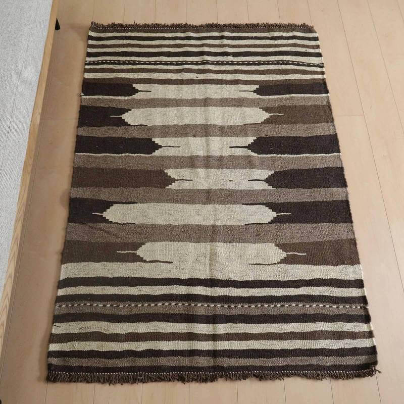 キリム・シラーズ／カシュガイ 153×118cm（KM-819）は、茶色とベージュのストライプが織りなす模様が特徴のギャッベラグが、ナチュラルな木目の床に敷かれている。