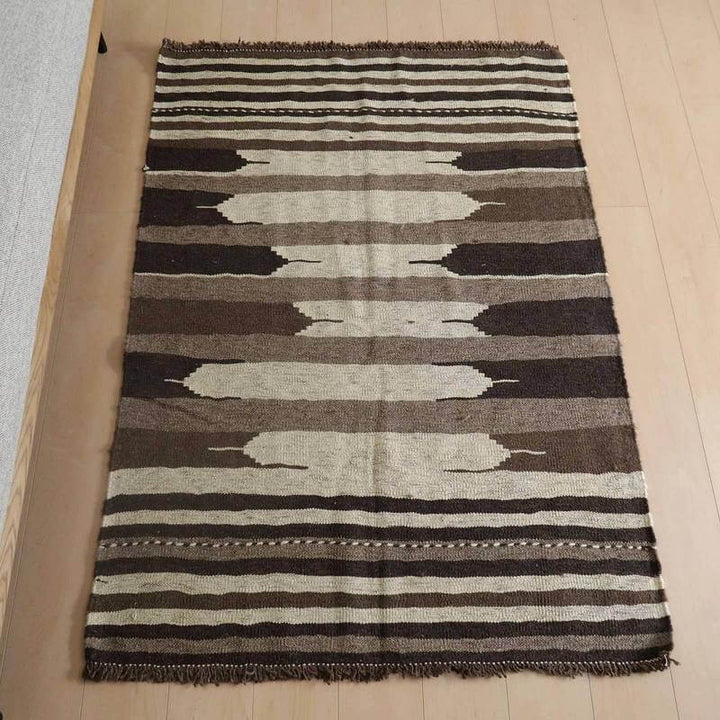 キリム・シラーズ／カシュガイ 153×118cm（KM-819）は、茶色とベージュのストライプが織りなす模様が特徴のギャッベラグが、ナチュラルな木目の床に敷かれている。
