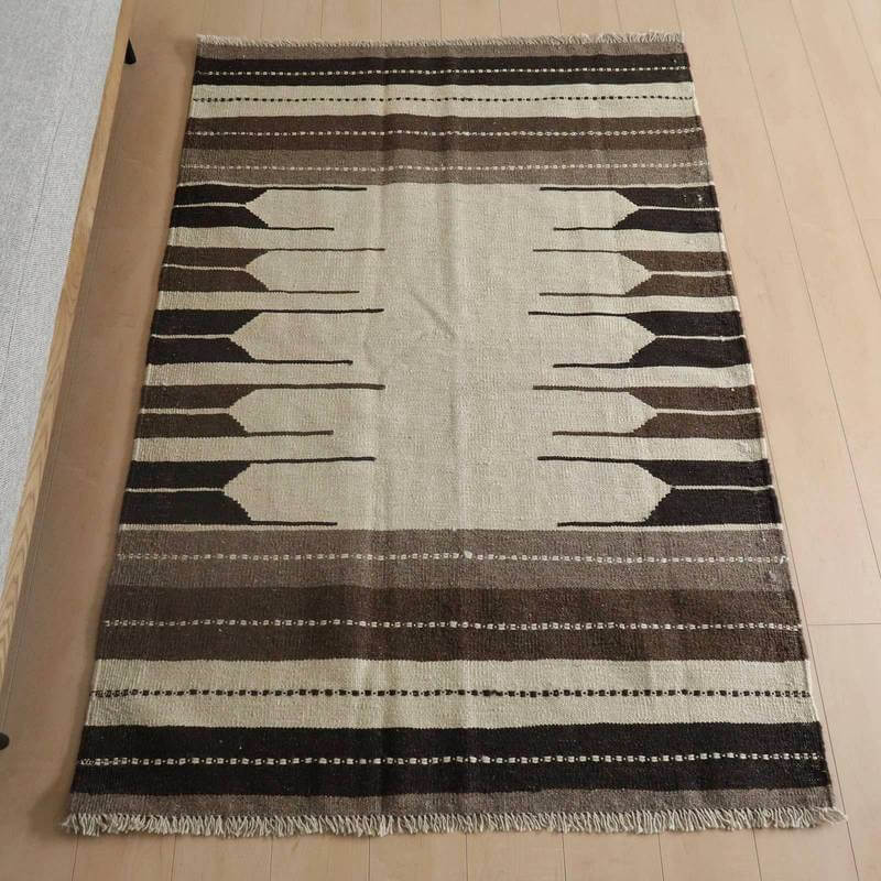 キリム・シラーズ／カシュガイ 178×122cm（KM-822）は、ブラウンとベージュの織り模様が美しいギャッベの手織りウールラグが床に敷かれている。
