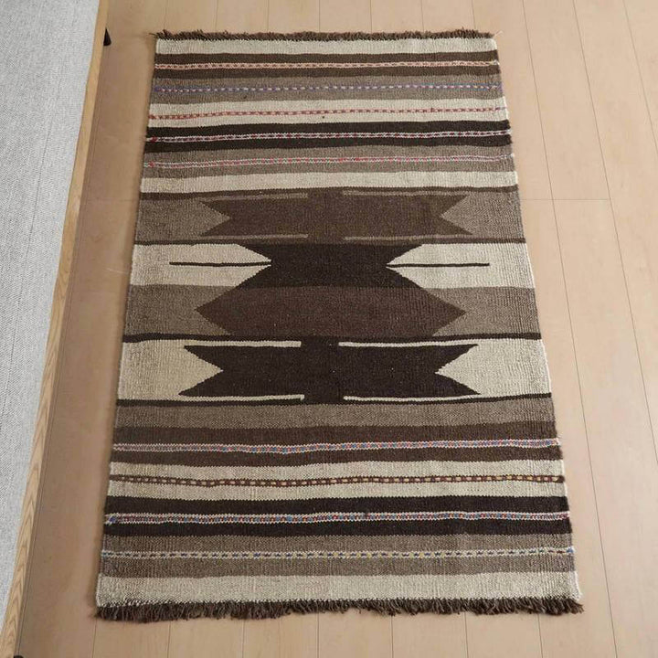 キリム・シラーズ／カシュガイ 151×98cm（KM-824）は、ウール素材で丁寧に手織りされたギャッベラグが床の上に美しく敷かれ、落ち着いたブラウンの模様が際立っている。