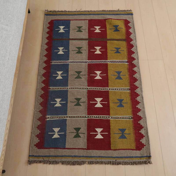 キリム・シラーズ／カシュガイ 153×97cm（KM-855）は、カラフルな幾何学模様が特徴のギャッベラグが、リビングの木製フローリングに美しく敷かれている。