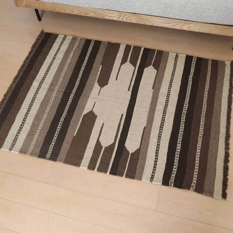 ブラウンとベージュのストライプ模様が印象的なギャッベの手織りウールラグが、シンプルな木製ベッドサイドに敷かれている。