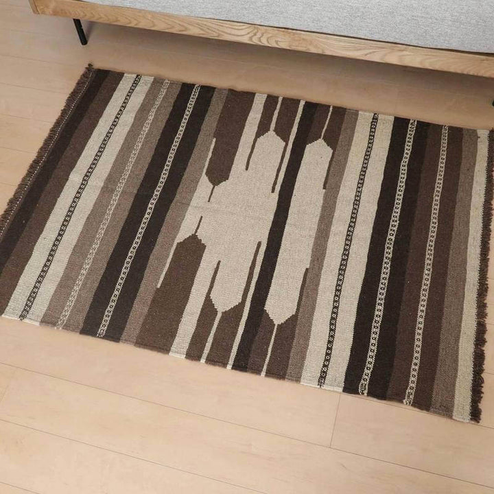 ブラウンとベージュのストライプ模様が印象的なギャッベの手織りウールラグが、シンプルな木製ベッドサイドに敷かれている。