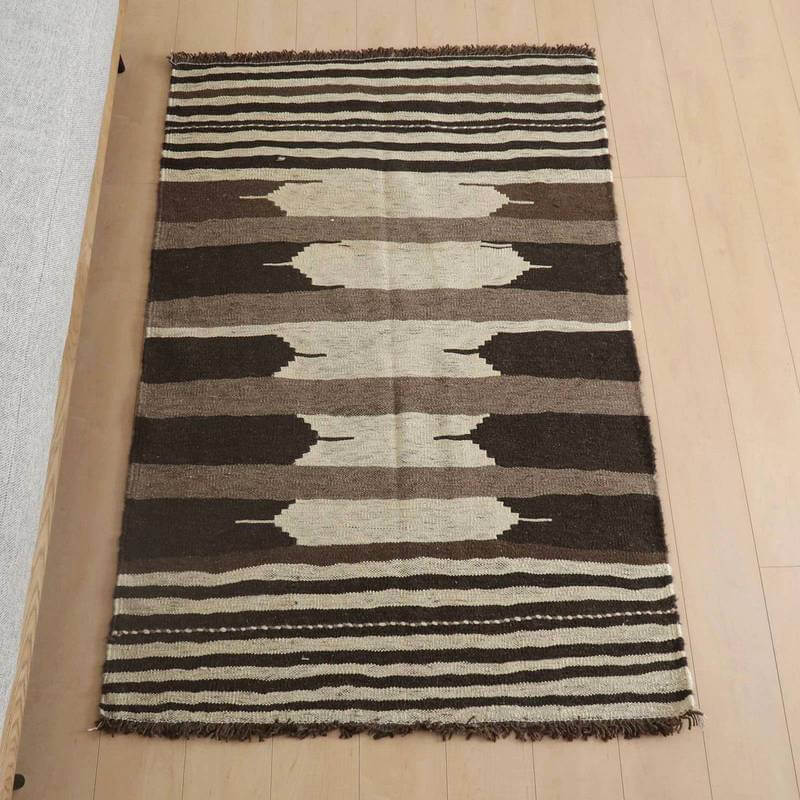 キリム・シラーズ／カシュガイ 154×104cm（KM-957）は、ブラウンとベージュのストライプが印象的なギャッベのラグが床に敷かれており、手織りのウール素材がリビングに落ち着いたぬくもりを添えている。