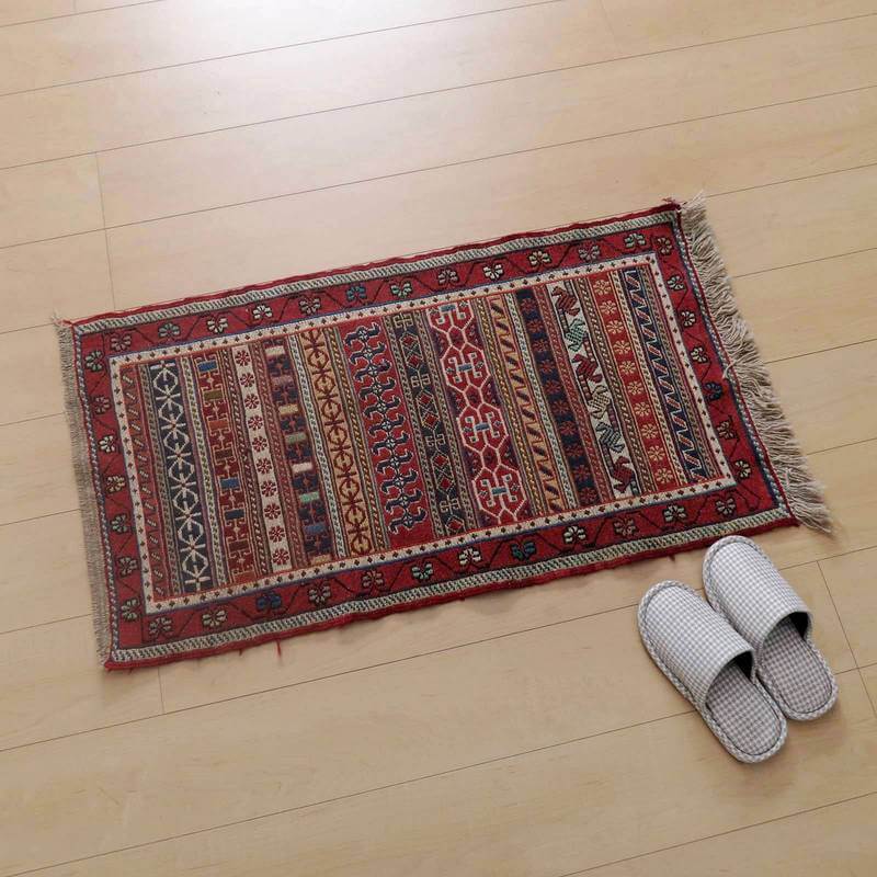 鮮やかな赤と複雑な模様が特徴のギャッベラグが、温かみのある木目床に美しく調和し、隣にはスリッパが並べられている。