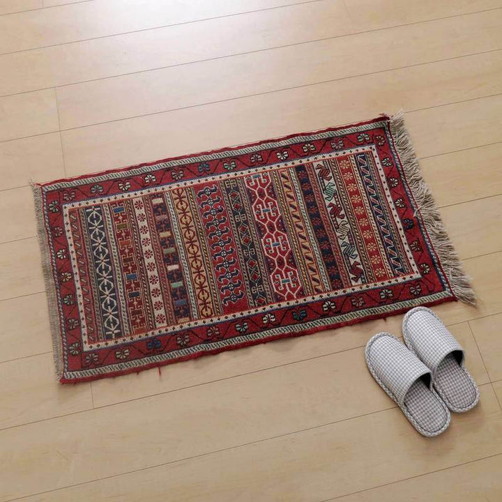 鮮やかな赤と複雑な模様が特徴のギャッベラグが、温かみのある木目床に美しく調和し、隣にはスリッパが並べられている。