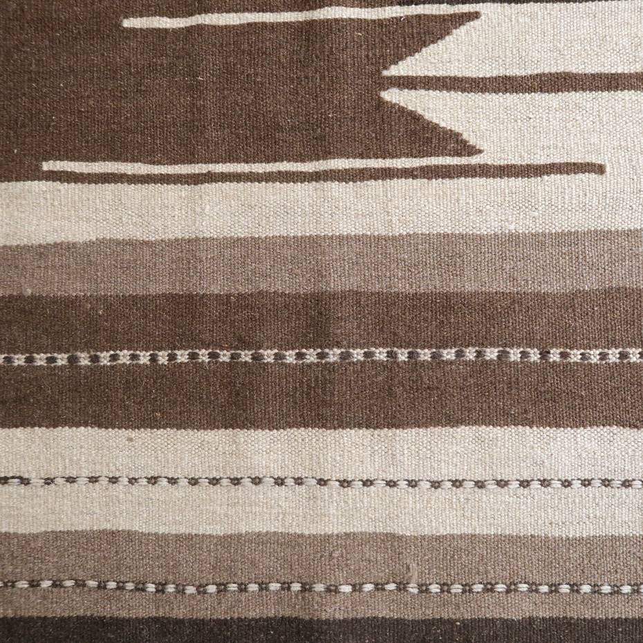 落ち着いたブラウンとベージュのストライプ模様が特徴の手織りギャッベラグが、ウール素材の温かみを感じさせるように床に広がっている。